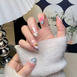 May include: Manicured nails featuring a beach-themed design with a soft pink base and light blue tips. The nails are decorated with small gold starfish and seashell charms, white pearls, and a three-dimensional pink flower accent on the index finger.