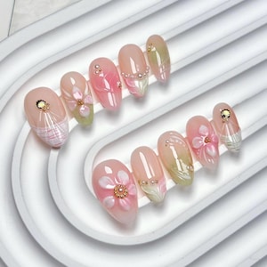 May include: A set of ten almond-shaped press-on nails featuring a translucent pink base with hand-painted floral designs, raised white petals, gold-toned metallic shell accents, and small embedded pearls arranged in various decorative patterns.