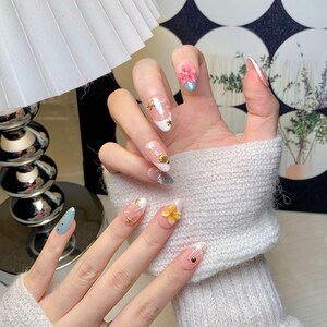 May include: Oval-shaped press-on nails featuring a light pink base with white tips, decorated with small gold starfish, a gold seashell, a pink flower, a yellow flower, and tiny white pearls. The nails have a glossy finish and a smooth texture.