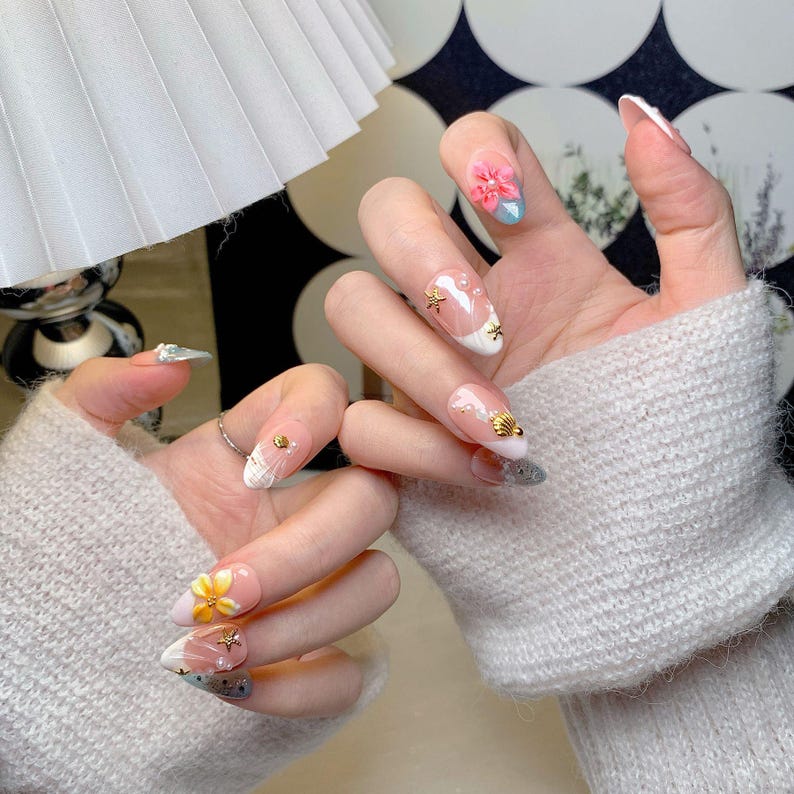 May include: Nails featuring a light pink base with white tips, adorned with three-dimensional floral accents in pink and yellow, small gold starfish charms, tiny white pearls, and subtle silver glitter gradients on the tips of several fingernails.