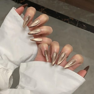May include: Long, almond-shaped press-on nails featuring a metallic gold cat-eye finish. The nails exhibit a reflective, shimmering texture that creates a distinct light-catching band across the centre of each nail, set against a white fabric sleeve.