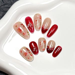 May include: A set of ten press-on nails featuring a glossy finish. The designs include a nude base with hand-painted pink cherry blossoms, solid deep red glitter, and red gradient tips. Some nails are accented with small white pearl embellishments.