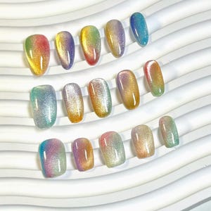 May include: An assortment of artificial nails in different shapes and sizes, displaying a spectrum of colours. The nails have a gradient effect, blending from one colour to another, including shades of red, yellow, green, blue, and purple. They are arranged on a white, wavy surface.