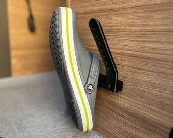 Op de afbeelding: Een zwarte schoenhanger gemonteerd op een houten paneel. Een grijze schoen met een groene streep hangt aan de hanger.
