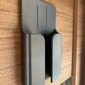 Op de afbeelding: Een zwarte 3D-geprinte telefoonhouder gemonteerd op een houten oppervlak. De houder heeft een gleuf voor een telefoon en een aparte gleuf voor oplaadkabels.