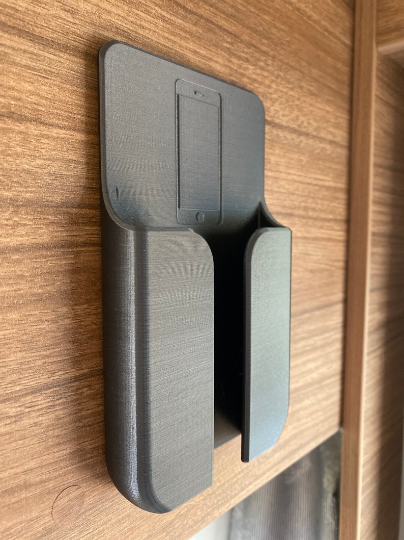 Op de afbeelding: Een zwarte 3D-geprinte telefoonhouder gemonteerd op een houten oppervlak. De houder heeft een gleuf voor een telefoon en een aparte gleuf voor oplaadkabels.