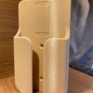 Op de afbeelding: Beigekleurige telefoonhouder van plastic met een gleuf voor een telefoon en een klein plankje aan de zijkant. De houder heeft twee gaten voor montage.