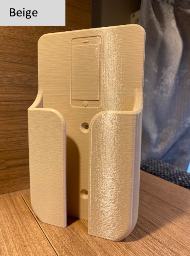 Op de afbeelding: Beigekleurige telefoonhouder van plastic met een gleuf voor een telefoon en een klein plankje aan de zijkant. De houder heeft twee gaten voor montage.