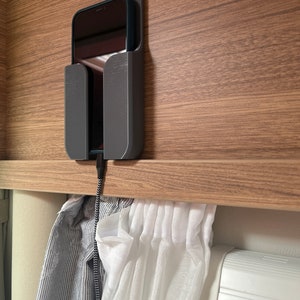 Op de afbeelding: Een zwarte telefoon die oplaadt in een grijze telefoonhouder die aan een houten oppervlak is bevestigd. De telefoon is verbonden met een zwarte oplaadkabel. Witte gordijnen hangen onder de telefoonhouder.