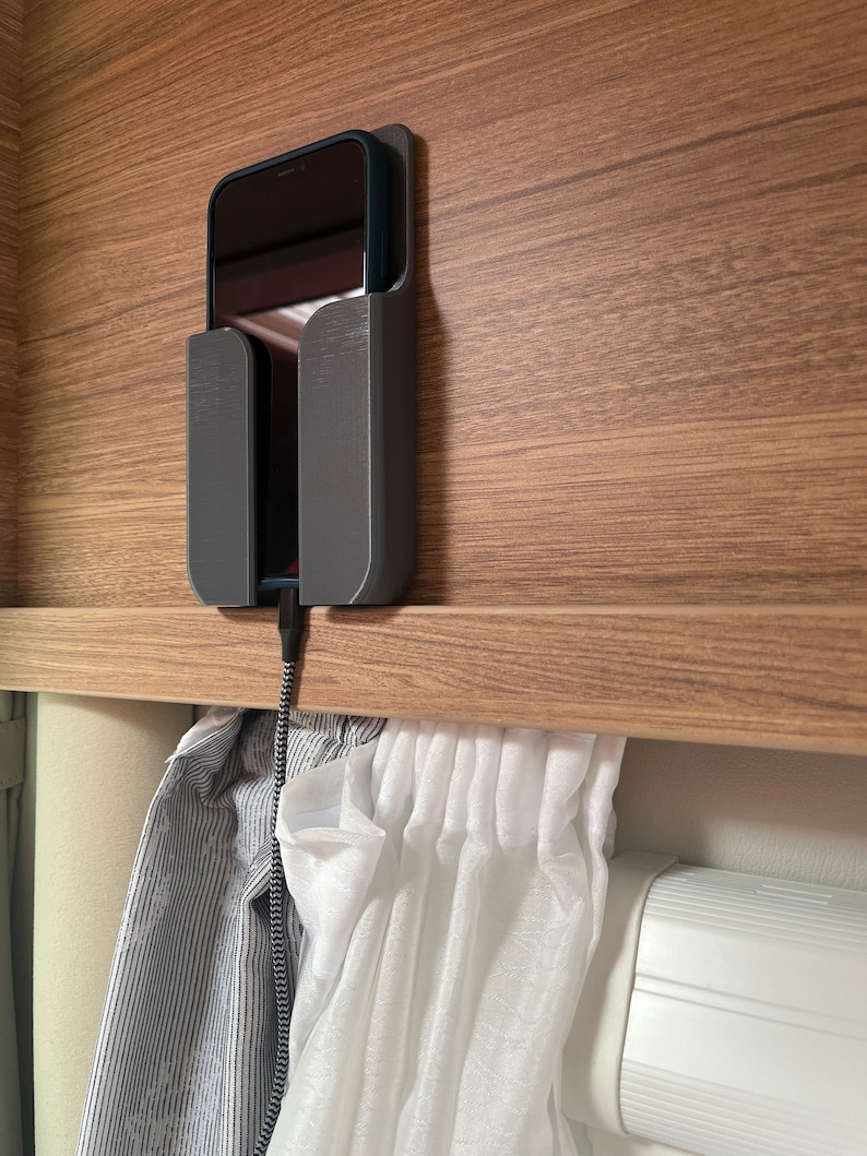 Op de afbeelding: Een zwarte telefoon die oplaadt in een grijze telefoonhouder die aan een houten oppervlak is bevestigd. De telefoon is verbonden met een zwarte oplaadkabel. Witte gordijnen hangen onder de telefoonhouder.