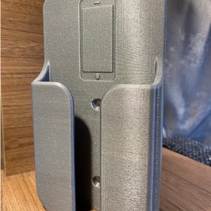 Op de afbeelding: Een zilveren metalen telefoonhouder met een uitsparing voor een telefoon. De houder heeft twee gaten aan de achterkant om te bevestigen.