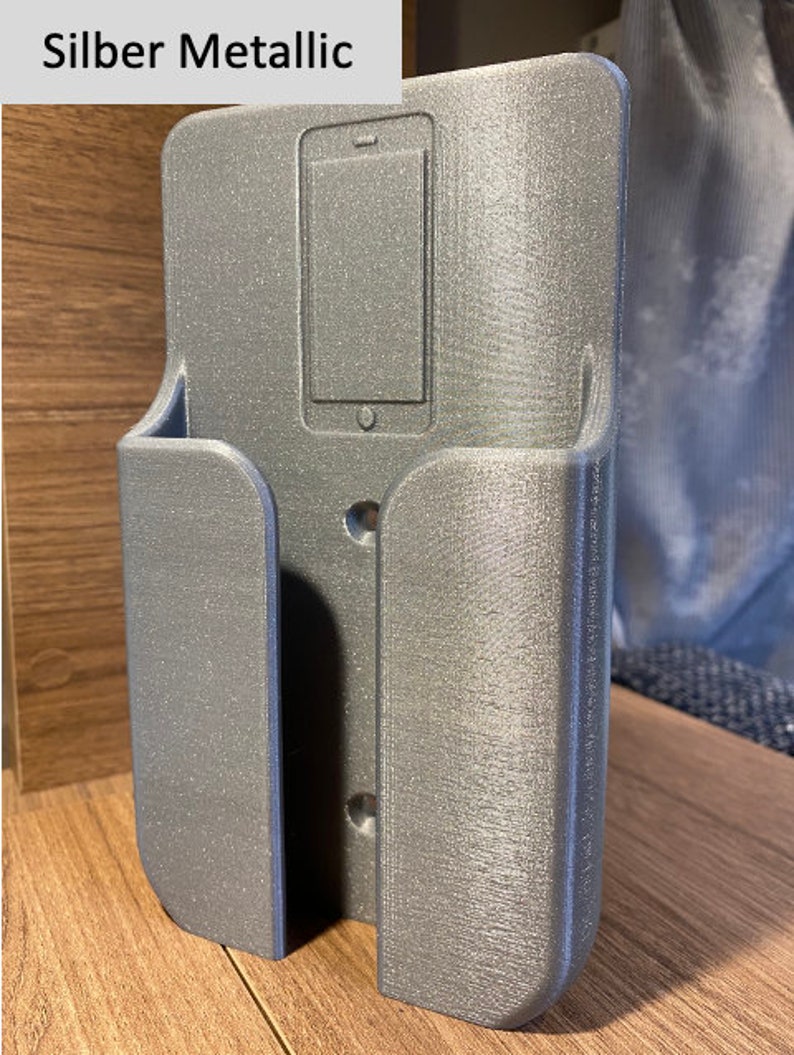 Op de afbeelding: Een zilveren metalen telefoonhouder met een uitsparing voor een telefoon. De houder heeft twee gaten aan de achterkant om te bevestigen.
