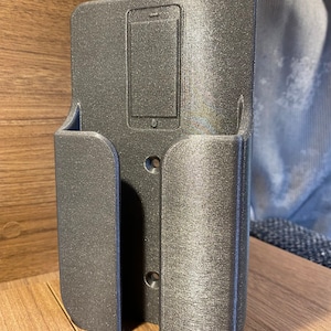 Op de afbeelding: Een zilvergrijze metalen telefoonhouder met een uitsparing voor een telefoon. De houder heeft een getextureerd oppervlak en is ontworpen om aan de muur te worden gemonteerd.