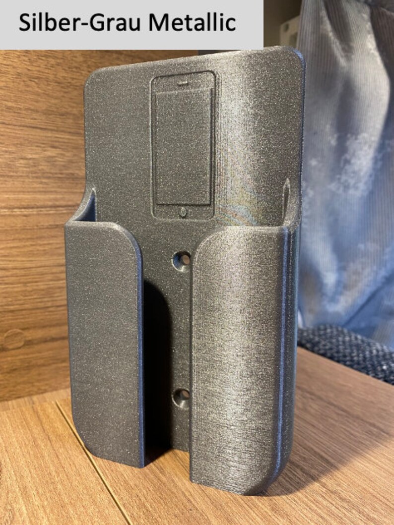 Op de afbeelding: Een zilvergrijze metalen telefoonhouder met een uitsparing voor een telefoon. De houder heeft een getextureerd oppervlak en is ontworpen om aan de muur te worden gemonteerd.