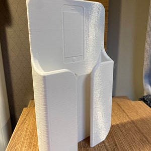 Op de afbeelding: Witte plastic telefoonhouder met een gleuf voor een telefoon en een aparte gleuf voor oplaadkabels. De houder is ontworpen om aan de muur te worden gemonteerd.