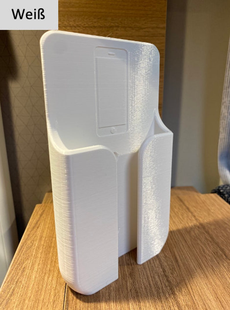 Op de afbeelding: Witte plastic telefoonhouder met een gleuf voor een telefoon en een aparte gleuf voor oplaadkabels. De houder is ontworpen om aan de muur te worden gemonteerd.