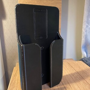 Op de afbeelding: Een zwarte plastic telefoonhouder met een gleuf voor een telefoon en een aparte gleuf voor oplaadkabels. De houder is aan een muur gemonteerd.