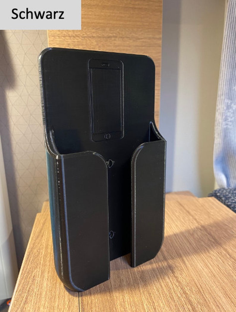 Op de afbeelding: Een zwarte plastic telefoonhouder met een gleuf voor een telefoon en een aparte gleuf voor oplaadkabels. De houder is aan een muur gemonteerd.