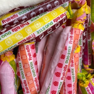 May include: A close-up of a pile of pink and white Fruit-tella Duo Stix candy. The candy is wrapped in colorful wrappers with the brand name and logo printed on them. The wrappers feature a variety of designs, including strawberries, lemons, and apples.