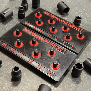 May include: Two black 3/8 shallow impact socket organizers with red lettering and accents. The trays feature labeled posts for sizes 7 through 19. Several individual black impact sockets are scattered around the organizers on the surface.