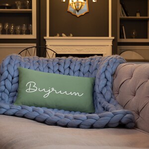 May include: A rectangular sage green throw pillow featuring the white cursive text "Buyrum" centred on the front, resting atop a light blue chunky knit blanket with thick, oversized yarn loops and a soft, textured appearance.