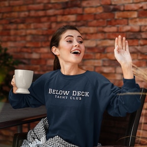 Op de afbeelding: Een vrouw draagt een donkerblauw sweatshirt met de tekst "BELOW DECK YACHT CLUB" in wit. Ze zit op een stoel met een deken en houdt een witte mok vast.