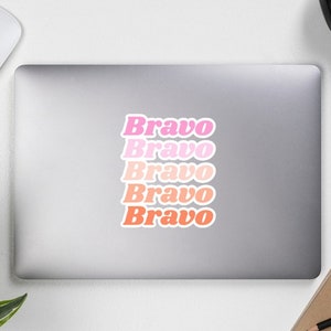 May include: A sticker with the word "Bravo" repeated five times in a gradient of pink and orange hues. The sticker is on a gray laptop, with a white mouse, headphones, and a notebook visible.