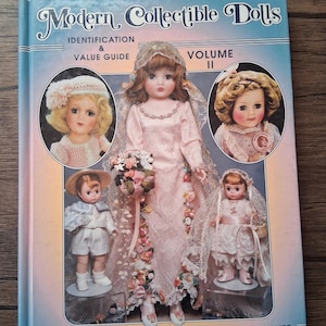 May include: A book titled "Modern Collectible Dolls, Identification & Value Guide, Volume II" featuring images of vintage dolls. The cover has a blue border and showcases several dolls in various outfits, including a bride and a young boy.