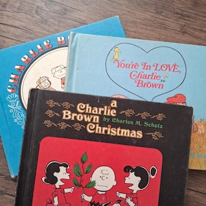 May include: Three vintage "Charlie Brown" books are stacked on a wooden surface. The books have blue and black covers with illustrations of the Peanuts characters. The titles include "Charlie Brown Christmas" and "You're in Love, Charlie Brown."