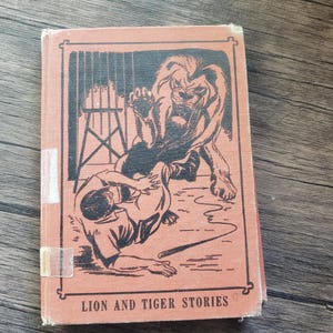 May include: Vintage book titled "Lion and Tiger Stories" with a cover illustration of a lion attacking a person. The cover is a faded orange color with black line art. The book is slightly worn.