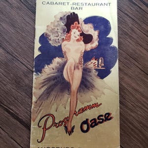 May include: Vintage cabaret restaurant bar menu. The menu features a stylized illustration of a woman with red hair and a large hat. The text on the menu reads "Programm Oase" and "Augsburg Tel. 36326". The background is gold.