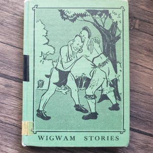 May include: A vintage book titled "Wigwam Stories" with a green cover. The cover features a black and white illustration of two figures under a tree. The book has a black spine and a yellowed label.