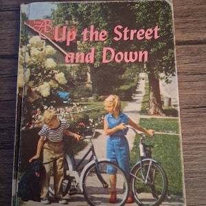 May include: Vintage book cover titled "Up the Street and Down." The cover features a colorful illustration of two children with bicycles and a black dog on a tree-lined street. The title is in pink letters.