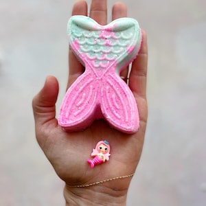 K&ouml;nnte beinhalten: Eine rosa und t&uuml;rkis farbene Badewannenbombe in Form einer Meerjungfrauenflosse mit Glitzerakzenten, gehalten in einer Hand. Eine kleine Plastik-Meerjungfrauenfigur befindet sich ebenfalls in der Hand. Die Badewannenbombe ist leuchtend rosa mit t&uuml;rkisfarbenen Akzenten.
