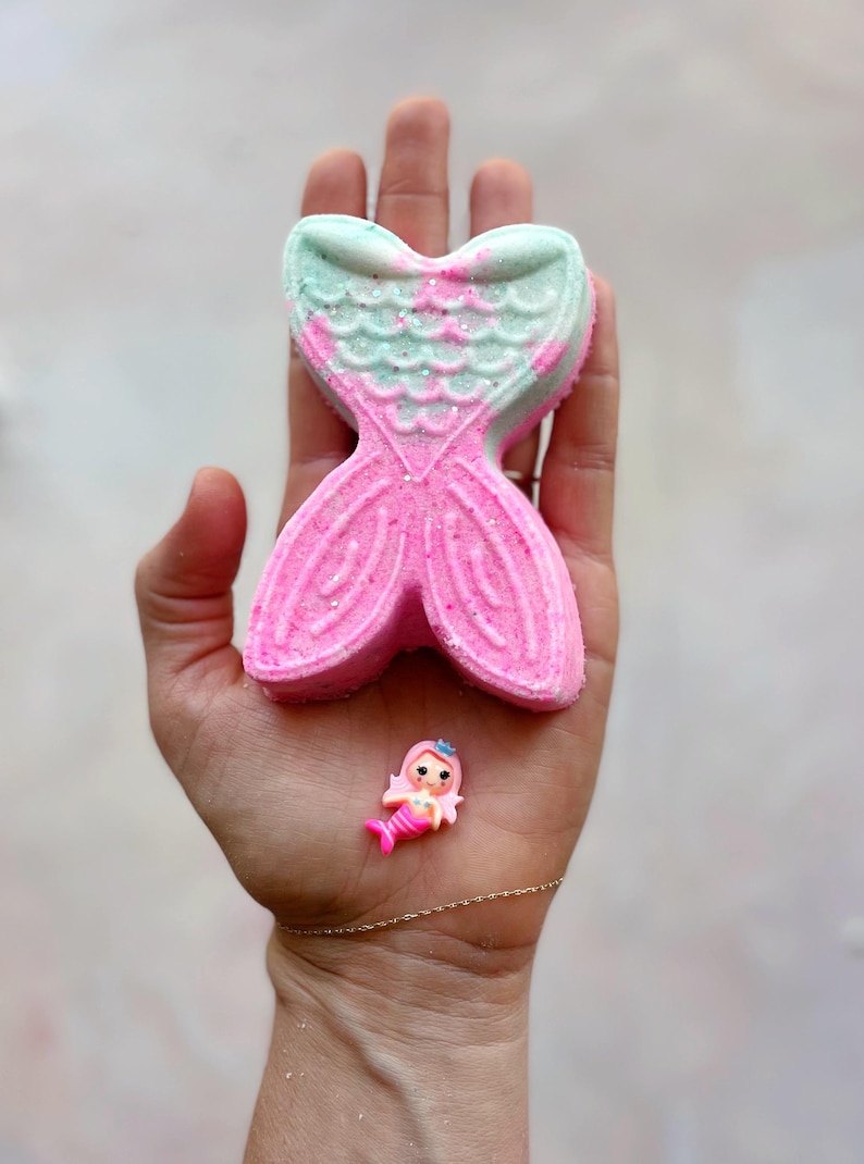 K&ouml;nnte beinhalten: Eine rosa und t&uuml;rkis farbene Badewannenbombe in Form einer Meerjungfrauenflosse mit Glitzerakzenten, gehalten in einer Hand. Eine kleine Plastik-Meerjungfrauenfigur befindet sich ebenfalls in der Hand. Die Badewannenbombe ist leuchtend rosa mit t&uuml;rkisfarbenen Akzenten.