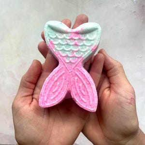 K&ouml;nnte beinhalten: Eine rosa und t&uuml;rkisfarbene Badewannenbombe in Form einer Meerjungfrauenflosse. Die Badewannenbombe wird in offenen H&auml;nden gehalten und hat ein strukturiertes Design. Der Schwanz ist rosa und die Oberseite ist t&uuml;rkis mit rosa Akzenten. Die Badewannenbombe ist so konzipiert, dass sie sich in Wasser aufl&ouml;st.