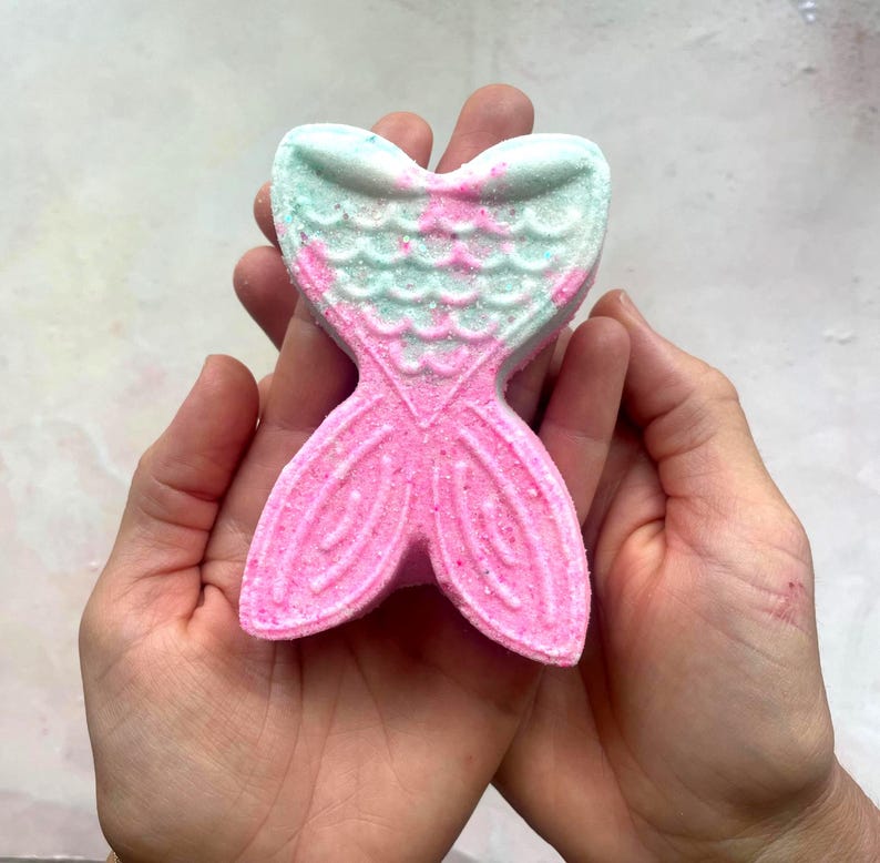 K&ouml;nnte beinhalten: Eine rosa und t&uuml;rkisfarbene Badewannenbombe in Form einer Meerjungfrauenflosse. Die Badewannenbombe wird in offenen H&auml;nden gehalten und hat ein strukturiertes Design. Der Schwanz ist rosa und die Oberseite ist t&uuml;rkis mit rosa Akzenten. Die Badewannenbombe ist so konzipiert, dass sie sich in Wasser aufl&ouml;st.