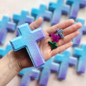 May include: A collection of cross-shaped items in shades of blue and purple. One large cross is held in a hand, with smaller crosses in various colors also visible. The crosses have a gradient effect and a sparkly finish.