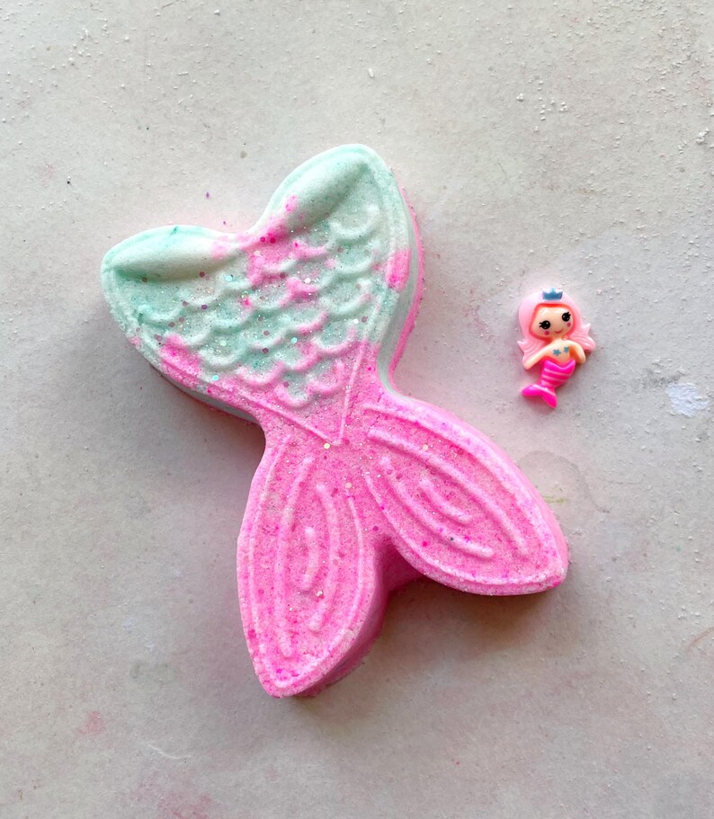 K&ouml;nnte beinhalten: Eine rosa und t&uuml;rkisfarbene Badewannenbombe in Form einer Meerjungfrauenflosse mit Glitzerakzenten. Neben der Badewannenbombe befindet sich eine kleine, rosa Meerjungfrauenfigur mit einer blauen Krone. Die Badewannenbombe hat ein strukturiertes, schuppenartiges Muster.