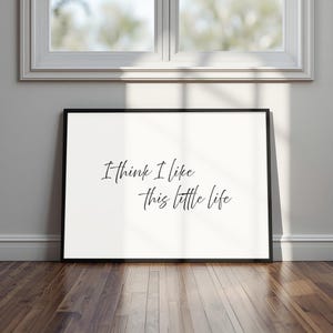 May include: A framed print with the handwritten text "I think I like this little life". The black frame surrounds a white background. The print is displayed on a wooden floor, near a window with a white frame.
