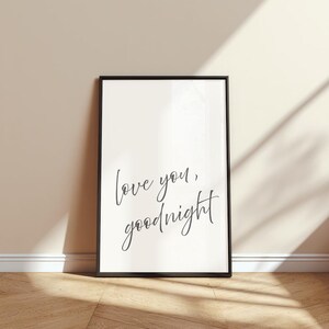 May include: A framed print with the words "love you, goodnight" in a cursive font. The black frame surrounds a white background. The print is leaning against a beige wall, with a wooden floor in the foreground.