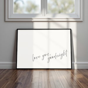 May include: A framed print with the words "love you, goodnight" in a handwritten script. The black frame surrounds a white background. The print is leaning against a wall on a wooden floor.