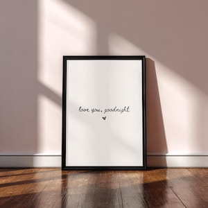 May include: A framed print with the words "love you, goodnight" in a handwritten style, with a small heart below the text. The black frame contrasts with the white background. The print is leaning against a wall on a wooden floor.