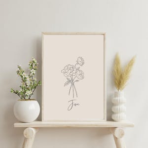 Op de afbeelding: Een ingelijste print met een minimalistische lijntekening van rozen en het woord "June" eronder. Het kunstwerk wordt tentoongesteld op een lichtgekleurde houten plank, met een witte vaas met bloemen en een decoratieve vaas met gedroogd gras.