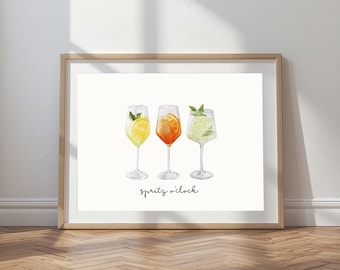 May include: A framed print featuring three watercolor cocktails in stemmed glasses. The drinks are yellow, orange, and green, garnished with citrus and herbs. The phrase "spritz o'clock" is written in cursive below the glasses. The frame is a light brown color.