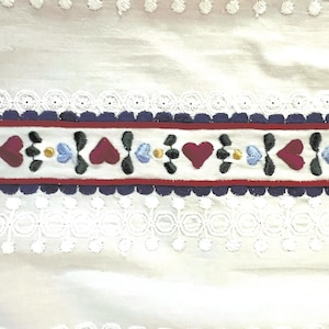 May include: White fabric with a red, blue, and black embroidered trim. The trim features a repeating pattern of hearts, leaves, and small flowers.