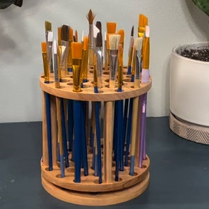 A wooden rotating paintbrush holder with a circular base and a cylindrical center. The holder has many holes to hold paintbrushes. The holder is filled with a variety of paintbrushes with different colored handles and bristles.