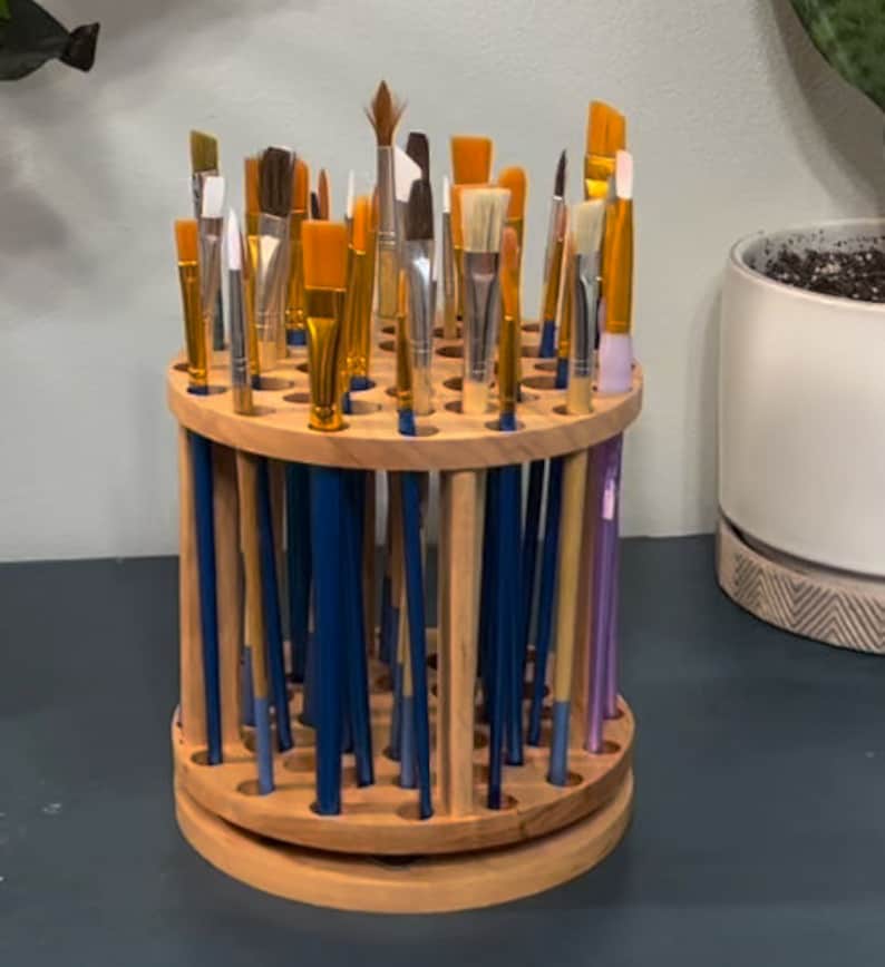 A wooden rotating paintbrush holder with a circular base and a cylindrical center. The holder has many holes to hold paintbrushes. The holder is filled with a variety of paintbrushes with different colored handles and bristles.