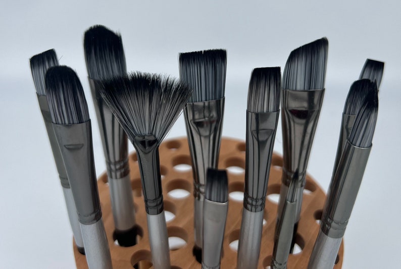 A wooden rotating paintbrush holder with a circular base and a cylindrical center. The holder has many holes to hold paintbrushes. The holder is filled with a variety of paintbrushes with different colored handles and bristles.