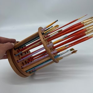 A wooden rotating paintbrush holder with a circular base and a cylindrical center. The holder has many holes to hold paintbrushes. The holder is filled with a variety of paintbrushes with different colored handles and bristles.